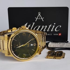 Puede incluir: Reloj de pulsera dorado con esfera verde oscuro, manecillas doradas y brazalete de eslabones dorados. El reloj se muestra con una tarjeta negra que dice "Atlantic" y una etiqueta de precio. También hay eslabones dorados adicionales.