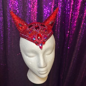 Könnte beinhalten: Ein rotes, mit Juwelen besetztes Kopfstück mit spitzen Ohren, das auf einem weißen Schaufensterpuppenkopf ausgestellt ist. Das Kopfstück ist mit roten Strasssteinen und größeren, facettierten Edelsteinen bedeckt. Der Hintergrund ist ein schimmernder violetter Stoff.