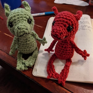May include: Two crocheted amigurumi figures, one green and one red, with black eyes and a toothy grin. The figures are sitting on a notebook.