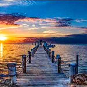 May include: A picturesque image of a wooden pier stretching out into the sea at sunset. The sky displays a gradient of blue, orange, and purple tones. The pier features wooden posts and a rope railing, leading to a small structure at the end.