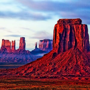 Op de afbeelding: Een panoramisch uitzicht op Monument Valley, een landschap van rode rotsformaties in Arizona, VS. De lucht is een levendig blauw met roze en paarse tinten. De rotsformaties zijn hoog en slank, met een grote, platte mesa op de voorgrond.