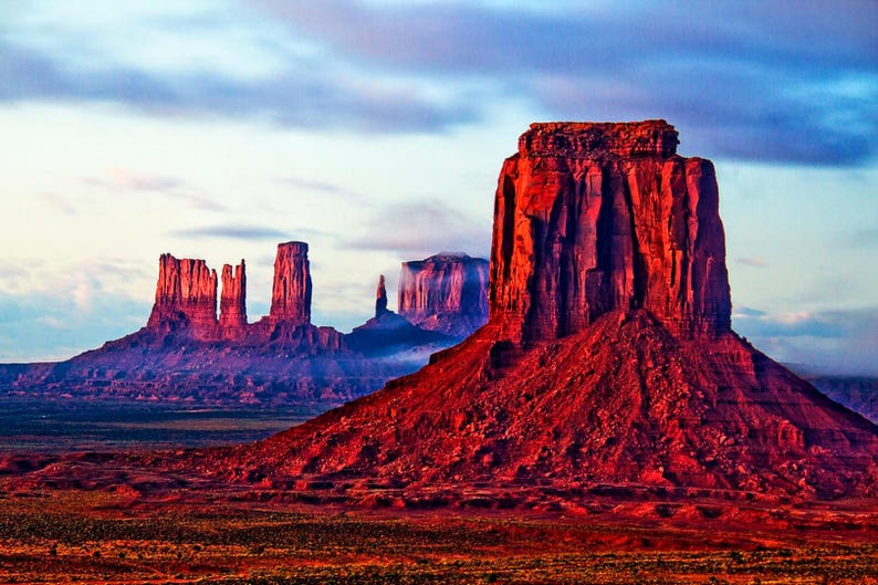 Op de afbeelding: Een panoramisch uitzicht op Monument Valley, een landschap van rode rotsformaties in Arizona, VS. De lucht is een levendig blauw met roze en paarse tinten. De rotsformaties zijn hoog en slank, met een grote, platte mesa op de voorgrond.