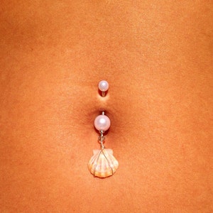 May include: A belly button ring with a pearl top and a dangling seashell charm. The ring is silver-toned with a large pearl in the center and a small pearl at the top. The seashell is beige and orange.