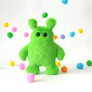 Puede incluir: Un juguete de peluche verde brillante con ojos grandes y una textura peluda. El juguete tiene un cuerpo redondeado y dos orejas pequeñas.