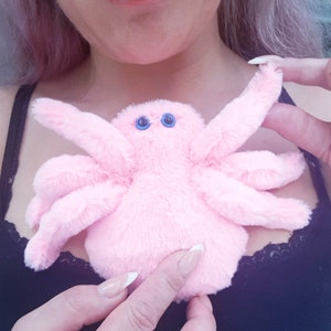 May include: A pink plush spider with blue eyes. The spider is being held by a person's hands.