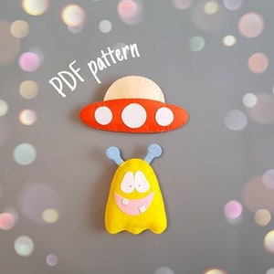 May include: PDF pattern for a felt alien toy with a big smile and a UFO. The alien is yellow with blue antennas and a pink mouth. The UFO is red with white circles.