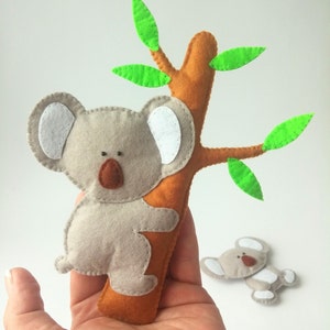 Pu&ograve; includere: Un koala di feltro grigio con orecchie bianche e occhi marroni sta arrampicandosi su un albero di feltro marrone con foglie di feltro verde.
