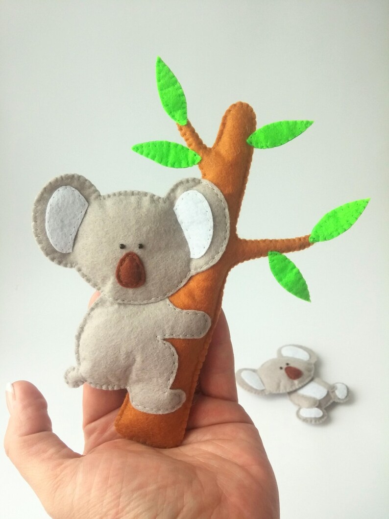 Pu&ograve; includere: Un koala di feltro grigio con orecchie bianche e occhi marroni sta arrampicandosi su un albero di feltro marrone con foglie di feltro verde.