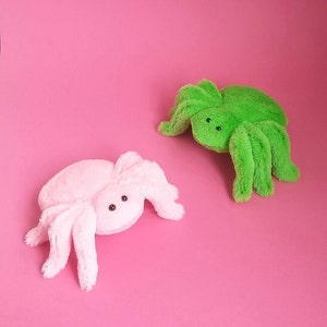 May include: Two plush spider toys, one pink and one green, on a pink background.