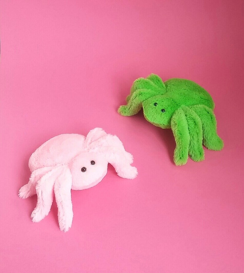 May include: Two plush spider toys, one pink and one green, on a pink background.