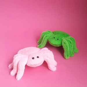 May include: Two plush spider toys, one pink and one green, on a pink background.