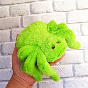 Pode incluir: Um brinquedo de aranha de pel&uacute;cia verde brilhante com olhos amarelos. A aranha est&aacute; sendo segurada na m&atilde;o de uma pessoa.
