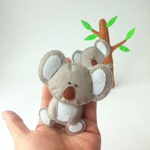 Pu&ograve; includere: Due orsi koala di feltro grigi con orecchie bianche e nasi marroni. Gli orsi koala sono seduti su una mano. L'orso koala in primo piano &egrave; tenuto da una mano. L'orso koala sullo sfondo &egrave; seduto su un albero di feltro marrone con foglie di feltro verde.