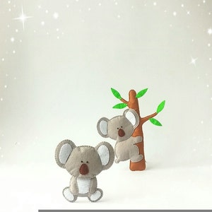 Pu&ograve; includere: Due orsi koala di feltro grigi, uno che scala un albero di feltro marrone con foglie di feltro verde, l'altro in piedi su una superficie bianca.