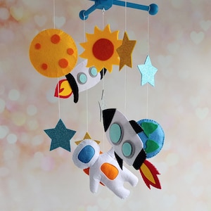 Puede incluir: Un móvil para bebé de fieltro con un marco de madera azul y cuerdas blancas. El móvil presenta un sol amarillo, un cohete azul y blanco, un astronauta blanco, una estrella azul, una estrella amarilla y un planeta azul y verde.