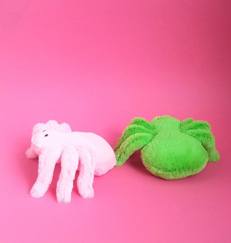 May include: Two plush spider toys, one pink and one green, on a pink background.