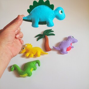 May include: Four felt dinosaur toys, including a blue stegosaurus, a yellow triceratops, a purple dinosaur, and a green brachiosaurus. The dinosaurs are all handmade and have a simple, childlike design.