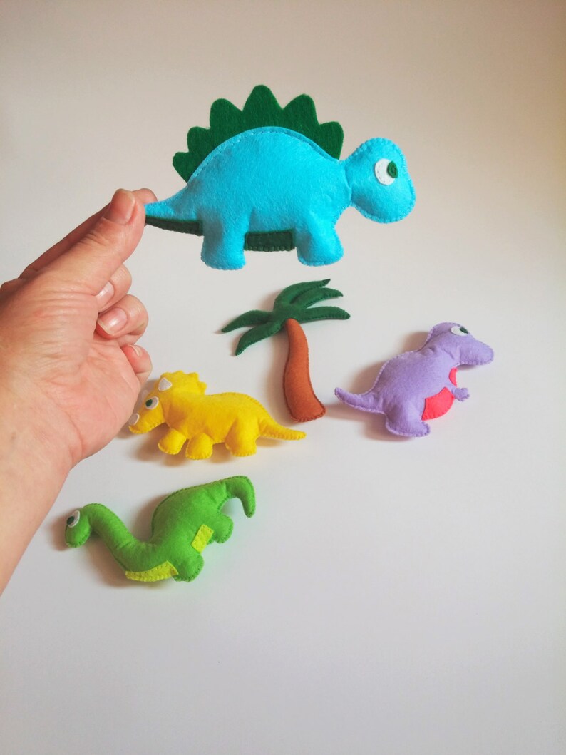 May include: Four felt dinosaur toys, including a blue stegosaurus, a yellow triceratops, a purple dinosaur, and a green brachiosaurus. The dinosaurs are all handmade and have a simple, childlike design.