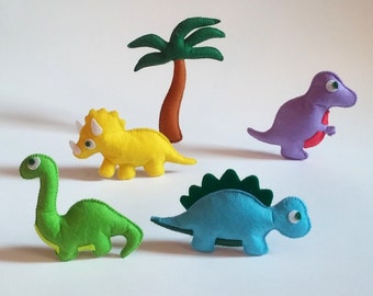 Può includere: Un set di cinque giocattoli di dinosauro in feltro, tra cui un dinosauro verde, un dinosauro blu con punte verdi, un triceratopo giallo, un dinosauro viola e una palma marrone.