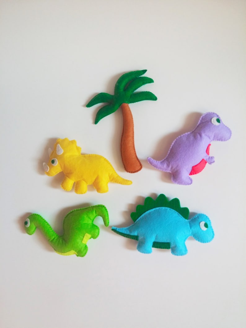 May include: Five felt dinosaur toys, including a green dinosaur, a blue dinosaur, a yellow dinosaur, a purple dinosaur, and a green palm tree.