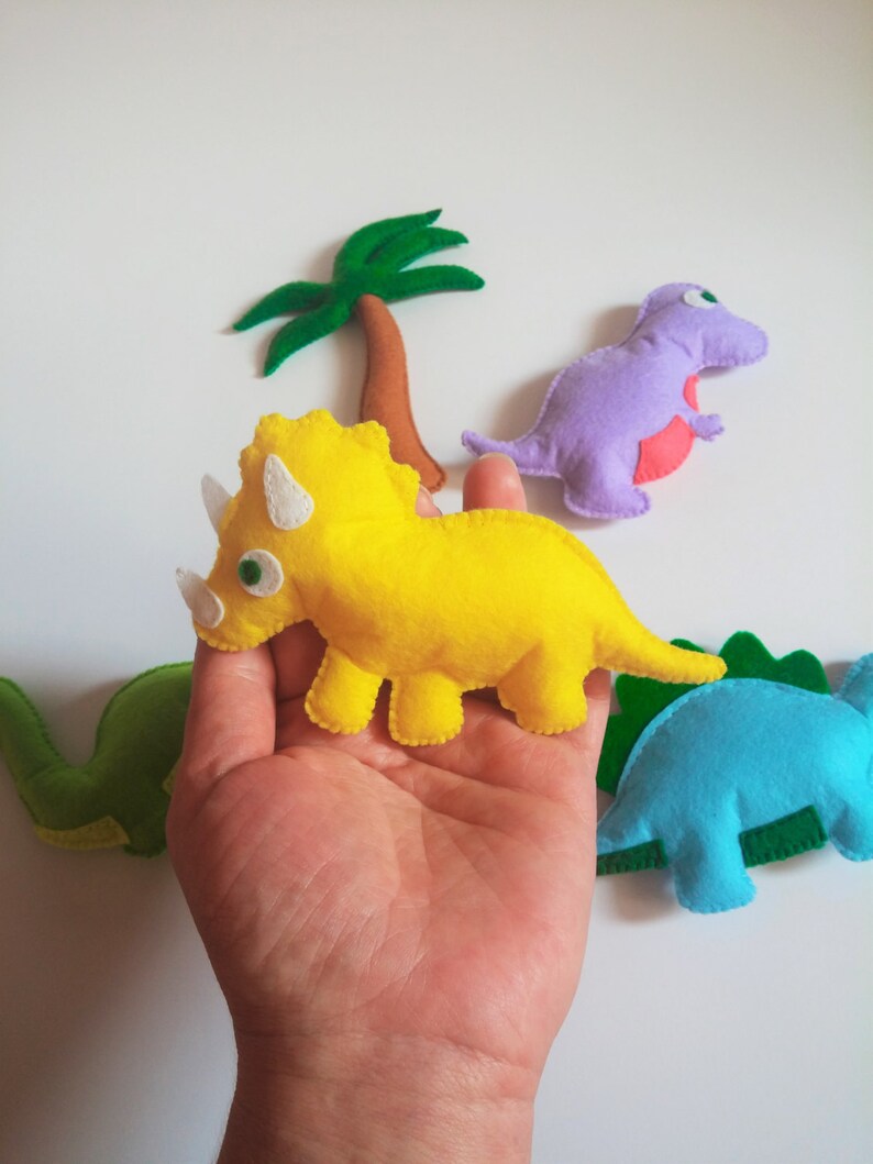 May include: A set of felt dinosaur toys, including a yellow triceratops, a purple tyrannosaurus rex, a blue stegosaurus, and a green brachiosaurus. The dinosaurs are all hand-sewn and have embroidered details.