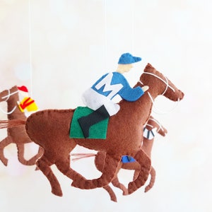 May include: A felt mobile with four brown horses, each with a jockey in a different colored uniform. The horses are in a running position, with their legs outstretched. The mobile is suspended from a red wooden bar.