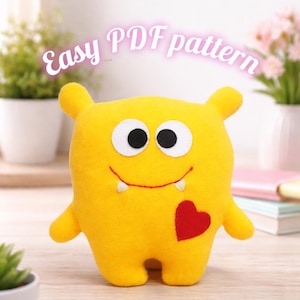 Puede incluir: Un monstruo de fieltro amarillo con ojos blancos, pupilas negras y un corazón rojo. El monstruo tiene brazos pequeños, orejas y dos pequeños colmillos. El texto "Easy PDF pattern" está encima del monstruo.