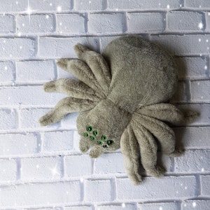 May include: A plush gray spider toy with green eyes. The spider is lying on a white brick background.