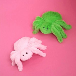 May include: Two plush spider toys, one pink and one green, on a pink background.
