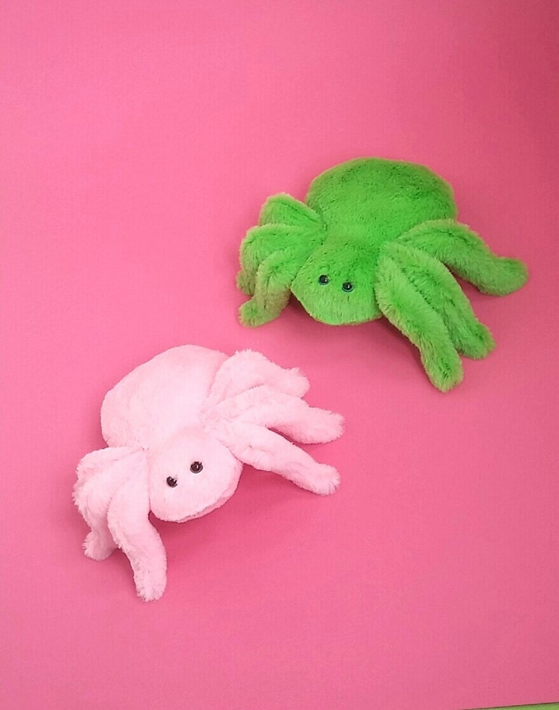 May include: Two plush spider toys, one pink and one green, on a pink background.