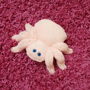 May include: A pink plush spider toy with blue eyes. The spider is lying on a pink fuzzy surface.