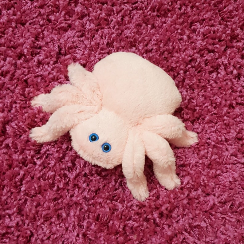 May include: A pink plush spider toy with blue eyes. The spider is lying on a pink fuzzy surface.