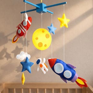 Op de afbeelding: Een baby mobile met ruimte thema vormen van vilt. Het bevat een blauwe raket, gele maan, sterren, wolken en een astronaut. De mobile hangt aan een blauw houten frame.