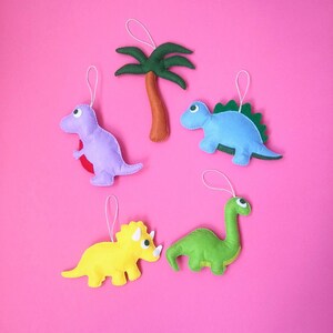 May include: Five felt ornaments, including a green dinosaur, a blue dinosaur, a yellow triceratops, a purple dinosaur, and a green palm tree. The ornaments are hanging on a pink background.