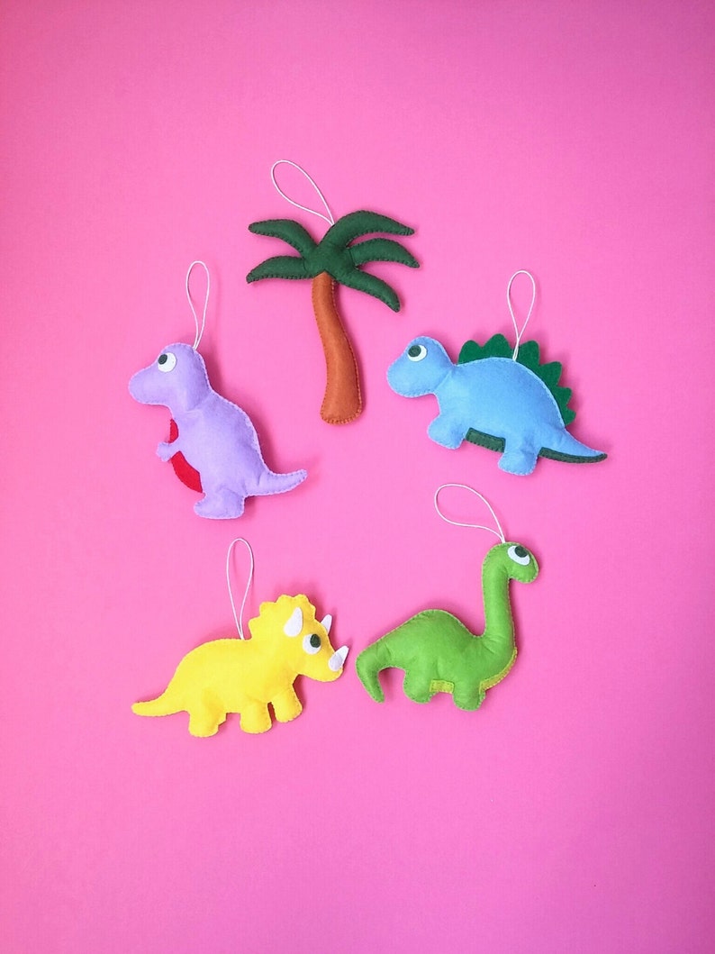 May include: Five felt ornaments, including a green dinosaur, a blue dinosaur, a yellow triceratops, a purple dinosaur, and a green palm tree. The ornaments are hanging on a pink background.