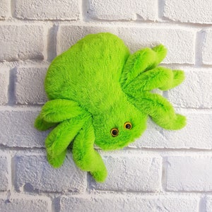 Pode incluir: Um brinquedo de aranha de pel&uacute;cia verde brilhante com grandes olhos amarelos. A aranha est&aacute; sentada em uma parede de tijolo branco.