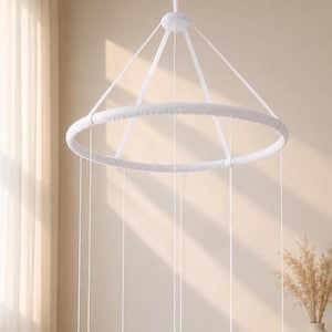 May include: A white circular mobile frame featuring a fabric-wrapped hoop suspended by four thin white cords that converge at a central top point. Multiple long, thin white strings hang vertically from the hoop, creating a minimalist structure.