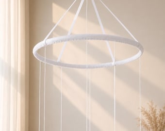 Op de afbeelding: Een wit cirkelvormig mobiel frame met een met stof beklede ring, opgehangen aan vier dunne witte koorden die samenkomen in een centraal punt aan de bovenkant. Meerdere lange, dunne witte draden hangen verticaal vanaf de ring naar beneden.