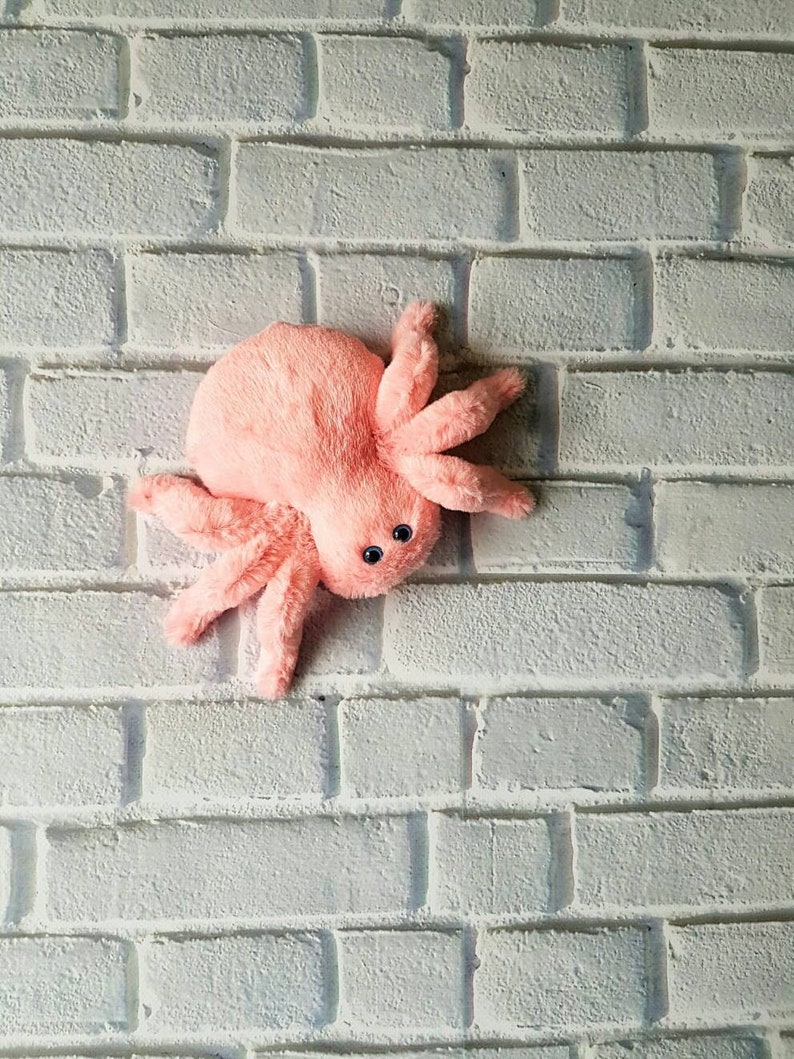 May include: A pink plush spider toy with black eyes. The spider is sitting on a white brick wall background.