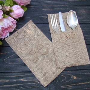 May include: Two burlap cutlery holders with a bow detail. One holder contains a fork, knife, and spoon. The holders are made of a natural tan burlap material. Pink flowers are in the background. The holders are on a dark wood surface.