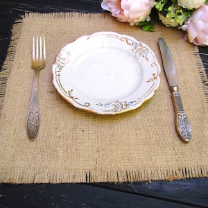 May include: A rustic table setting with a white plate with gold trim and blue accents, a fork, and a knife. The plate rests on a burlap placemat with frayed edges. Pink flowers are in the background.