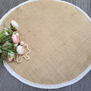 May include: A round, beige burlap table runner with a white lace trim. The runner is decorated with a bouquet of pink flowers and a pearl necklace.