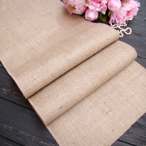 May include: A folded, beige burlap table runner. The fabric has a woven texture and is draped over a dark wooden surface. Pink peonies and a string of pearls are in the background, suggesting a rustic or farmhouse theme.