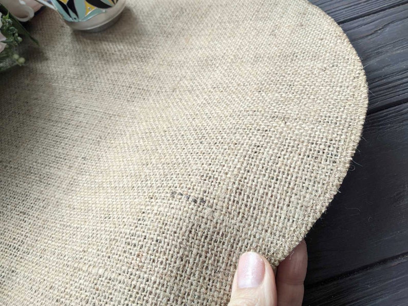 May include: A round, natural burlap placemat with a woven texture. The placemat is beige in color and has a slightly frayed edge.