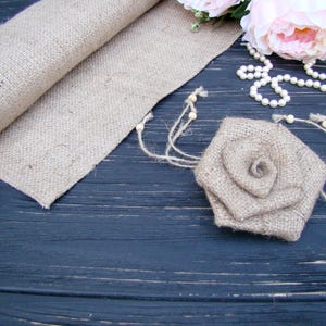 Pu&ograve; includere: Un runner in juta e una rosa in juta su una superficie in legno scuro. La rosa &egrave; fatta di juta e ha un aspetto rustico e naturale. Il runner &egrave; arrotolato e ha un aspetto rustico e naturale. C'&egrave; una collana di perle bianche sulla superficie.
