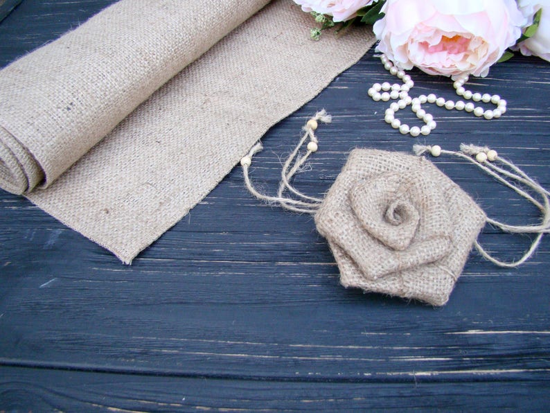 Pu&ograve; includere: Un runner in juta e una rosa in juta su una superficie in legno scuro. La rosa &egrave; fatta di juta e ha un aspetto rustico e naturale. Il runner &egrave; arrotolato e ha un aspetto rustico e naturale. C'&egrave; una collana di perle bianche sulla superficie.