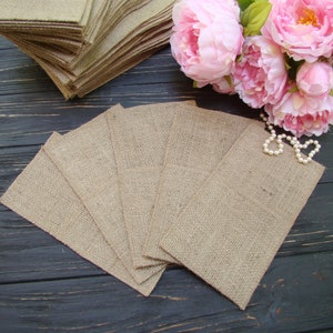 May include: A set of eight small burlap bags, folded and arranged in a fan shape on a black wooden surface. The bags are made of natural burlap fabric and have a rustic, handmade look.