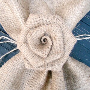 Pu&ograve; includere: Un fiore di juta con un centro marrone, legato con uno spago, su uno sfondo di legno blu scuro.