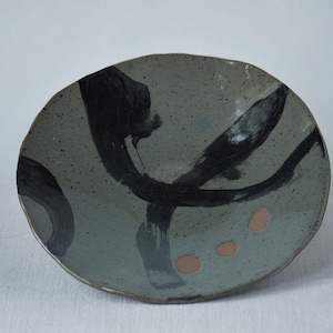 May include: A handmade ceramic plate with a gray-green glaze. The plate features bold black brushstrokes and three small, round, orange-brown accents. The plate has an organic, irregular shape, suggesting a unique, artisanal quality.