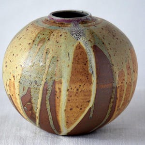 May include: A round ceramic vase with a small opening. The vase has a speckled, textured surface with a color palette of tan, brown, and green. Vertical streaks of brown and tan run down the sides, creating a unique design.